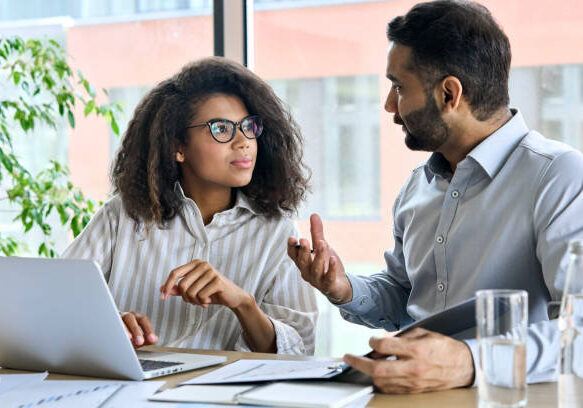 Businessman indian executive manager ceo talking to female African American coworker, using laptop. Diverse multicultural professional partners group discussing business plan at board room meeting.